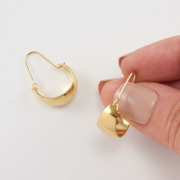 Fingers holding one of a pair of gold convex curved hoop earrings.