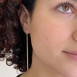 Close up front view of woman wearing pair of long gold earrings of thin cast feather on earwire.