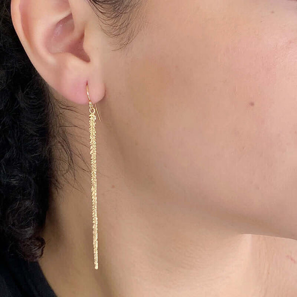 Close up side view of woman wearing pair of long gold earrings of thin cast feather on earwire.