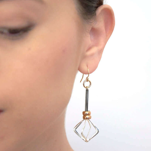 Close-up front view of woman wearing dark chain dangle earrings with cast silver rhombus.