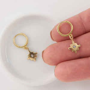Fingers holding one of a pair of gold native motif cross earrings on hoop clasp.