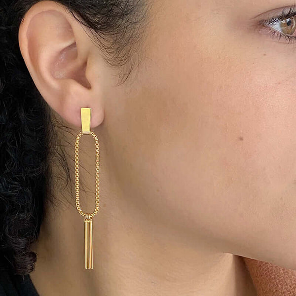 Close-up side view of woman wearing a pair of gold earrings, chain hoop with 3 hanging bars on posts.