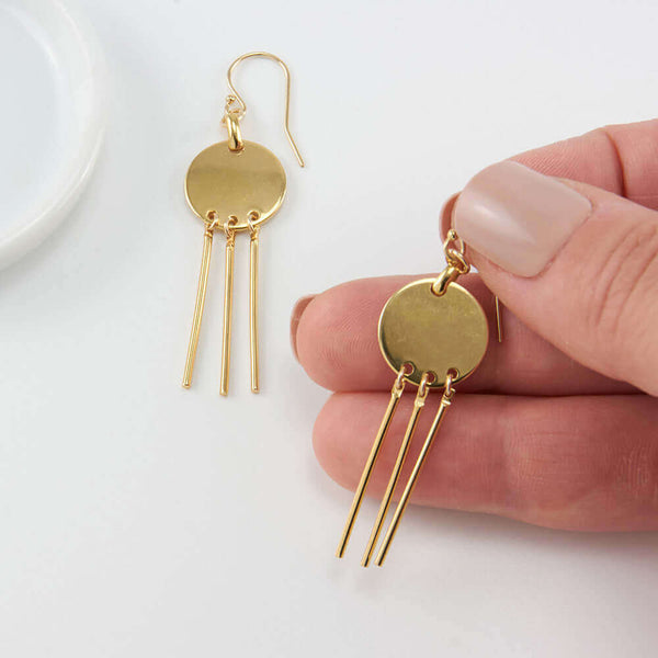 Fingers holding one of a pair of gold earrings, round disc with 3 hanging bars on earwire.