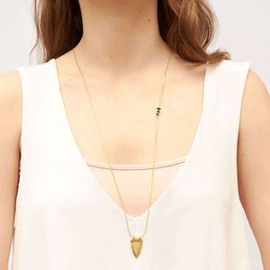 Woman wearing long gold necklace with shield shaped pendant, hangs down to mid torso.