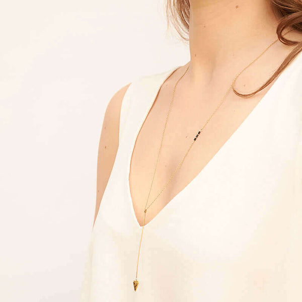 Woman wearing gold necklace, long arrow pendant, chain with black bead accents, shown from side angle.