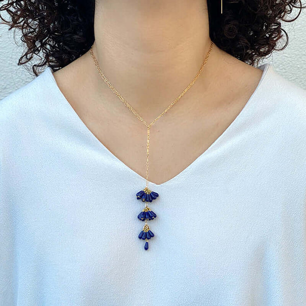 Woman wearing gold chain necklace with pendant of 3 bunches dark blue beads and single bead at end, worn short.