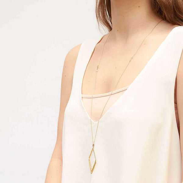 Woman wearing long gold necklace with rhombus shaped pendant, shown side angle.