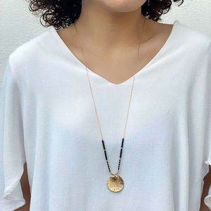 Woman wearing gold necklace with round medallion and black beads on either side, hanging long.