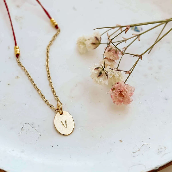 Close-up of delicate red thread necklace with gold chain and oval pendant on plate with small flowers.