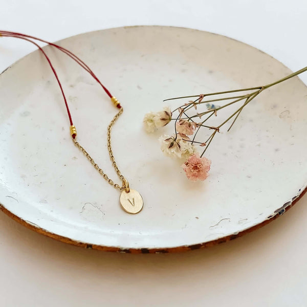 Close-up of delicate red thread necklace with gold chain and oval pendant on plate with small flowers.