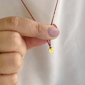Close-up of fingers holding delicate red thread necklace with gold accents and oval pendant.