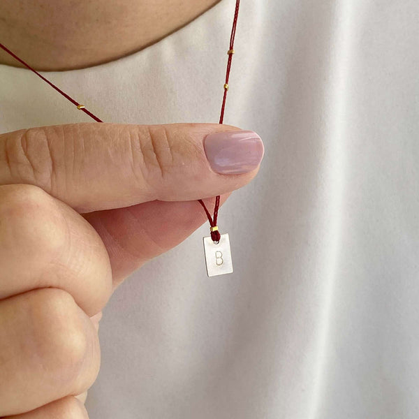Close-up of fingers holding delicate red thread necklace with gold accents and square pendant.