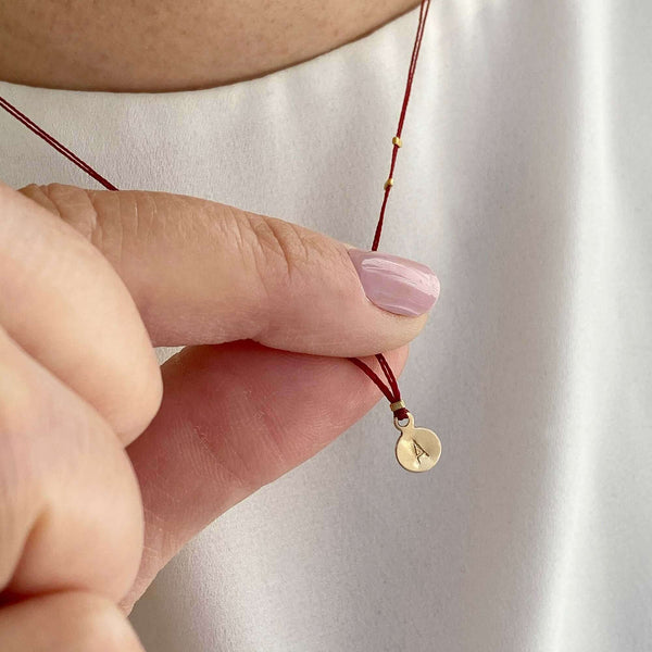 Close-up of fingers holding delicate red thread necklace with gold accents and round pendant.