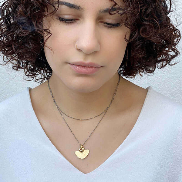 Woman wearing delicate dark chain necklace with tiny gold beads along chain and gold half-circle shaped pendant. worn doubled up.