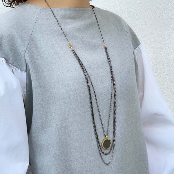 Woman wearing dark multi-chain necklace with round coin shaped etched brass pendant, worn long.