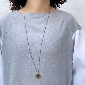 Woman wearing long dark chain necklace with etched coin shaped pendant.