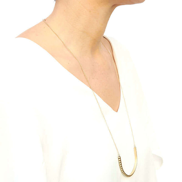 Woman wearing gold necklace with large curved beaded bar pendant, shown side angle.